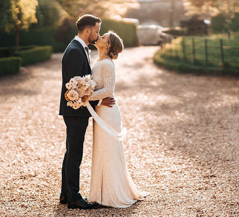 Bride in long sleeve floor length beaded wedding dress with rust, toffee roses and beige flowers kisses groom in three piece suit