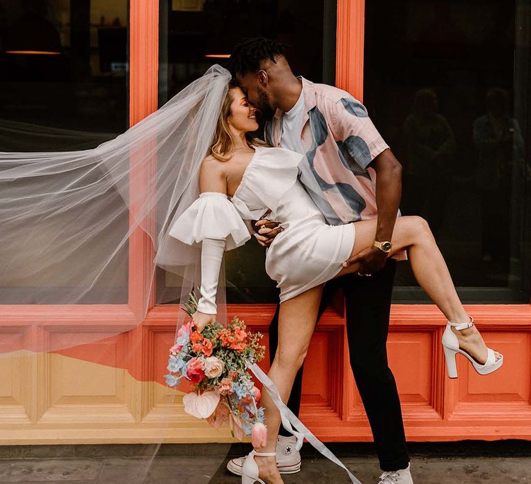 Groom in a patterned shirt leaning his bride over for a kiss in a short wedding dress and long veil at their stylish city elopement 