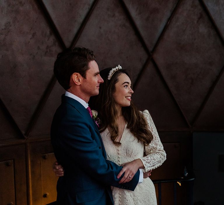 Smiling bride in lace Rixo dress and groom in blue suit take a look out at the view of London city and the Millennium Dome