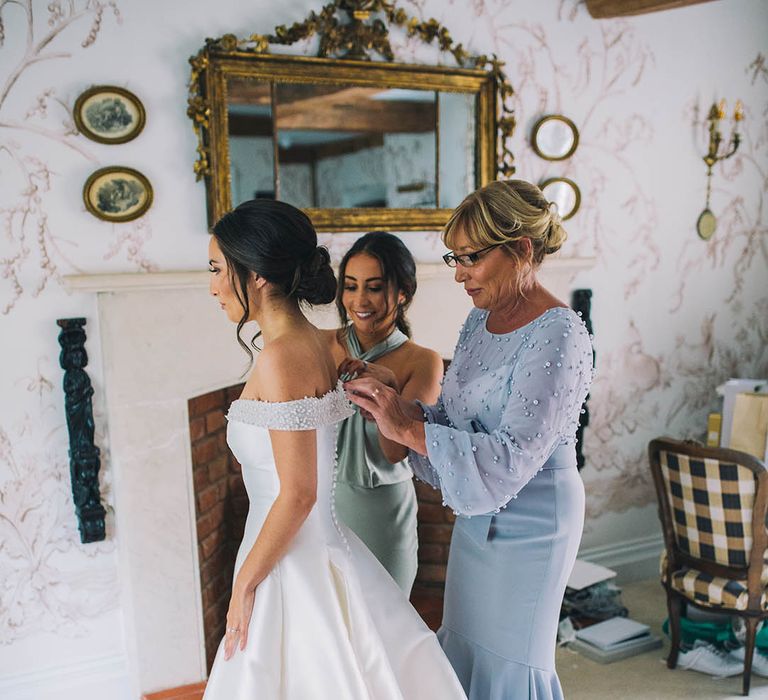 Mother of the bride in blue mermaid style dress and bridesmaid in mint green dress helps bride get into her wedding dress