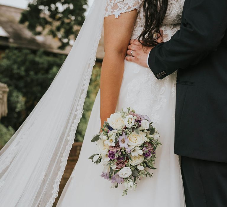 Bride's lace detailed wedding dress and veil with white and purple bouquet as she is held by the groom