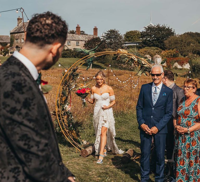 Bride in lace boho wedding dress and blues shoes walks through wooden moongate with dried flowers holding bright bouquet 