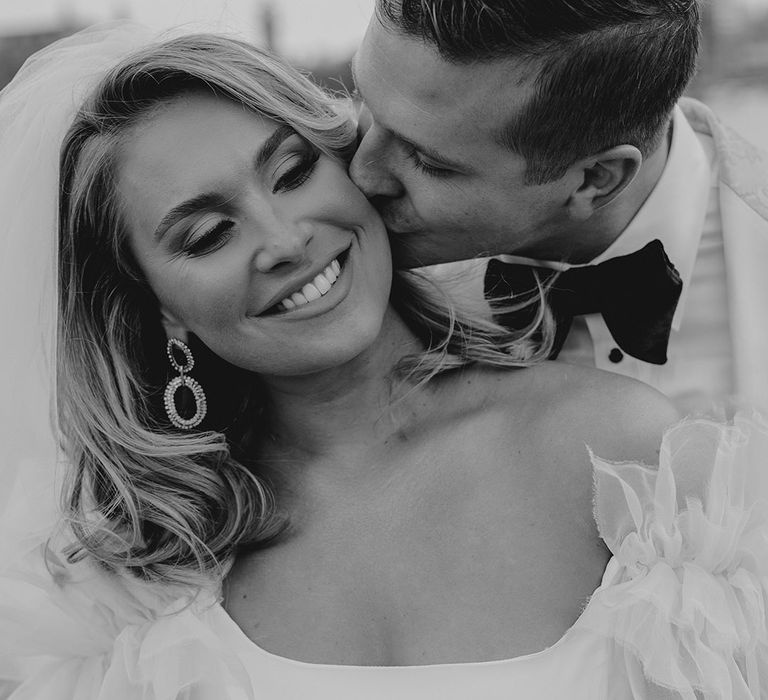 Bride with smoky eye makeup gets kissed on the cheek by groom in black tie