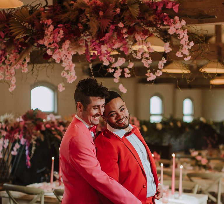 Groom in a pink suit and white boots embracing his partner in a red suit under a wooden beam decorated with cherry blossom and pink flowers 