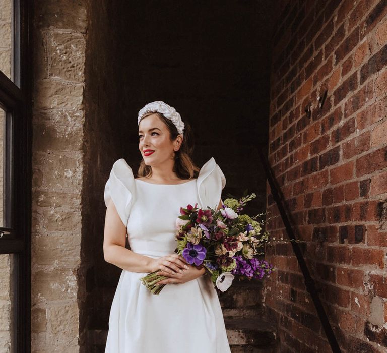 Bride in Jesus Peiro wedding dress with raised ruffle sleeves with white headband holding purple, burgundy and white bouquet 