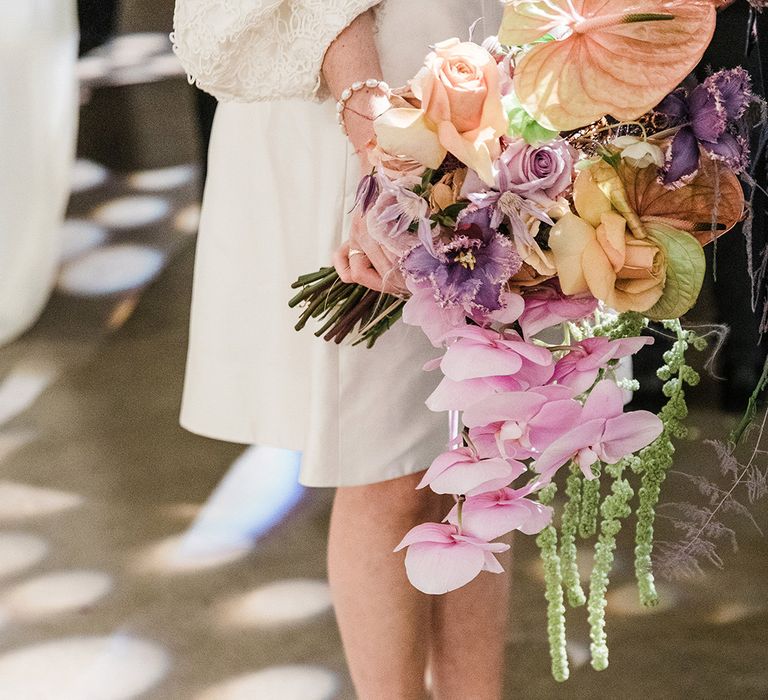 Bride holding orange and purple wedding bouquet with orchids and roses wearing short wedding dress and velvet platform block heels