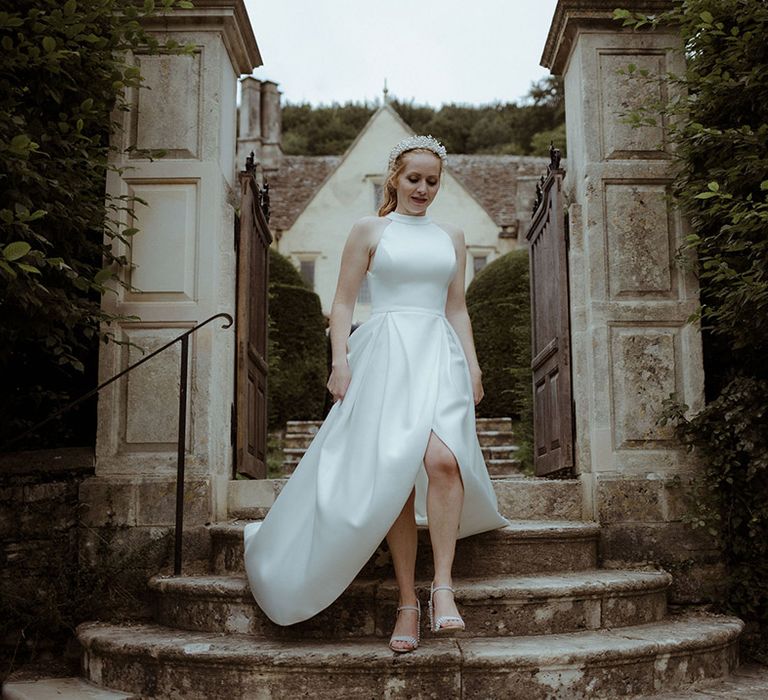 Bride in halter neck wedding dress with pleated skirts, a pearl crown and pearl embellished wedding shoes walks down some stairs 