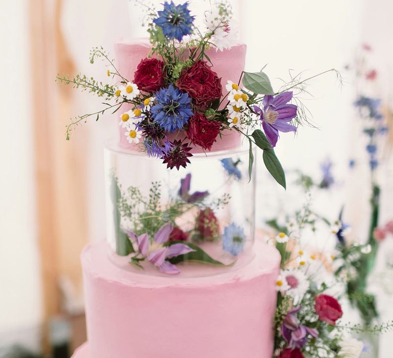 Four tier pink frosted wedding cake with plastic layer with purple wildflowers on wood slice cake stand