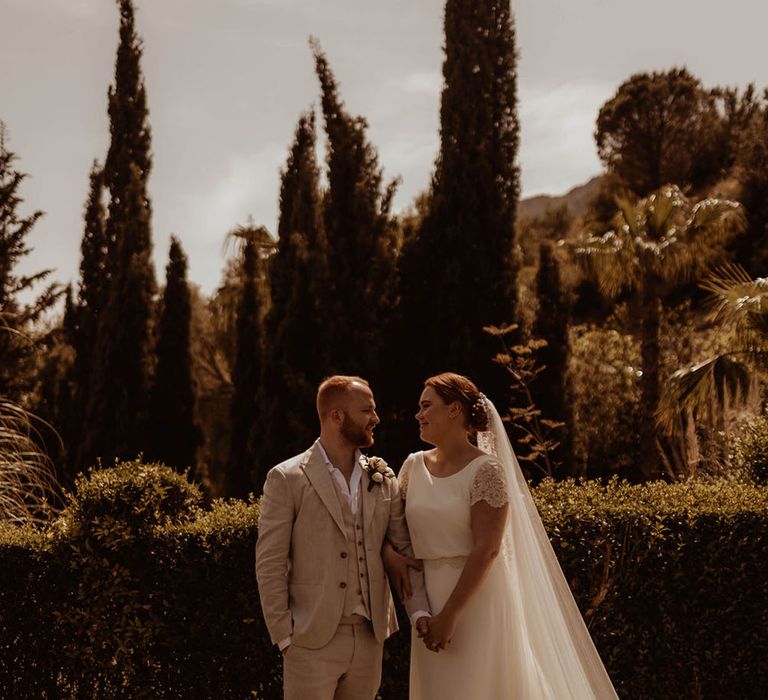 Bride & groom look lovingly at one another as groom wears three piece cream suit complete with relaxed open shirt 