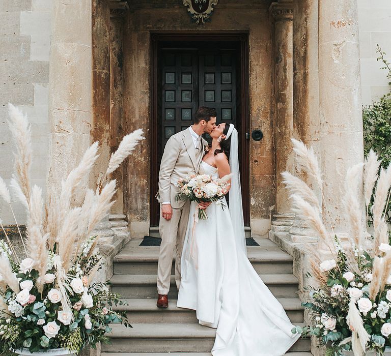 Bride and groom kiss at the entrance to Elmore Court with pampas grass floral arrangements 