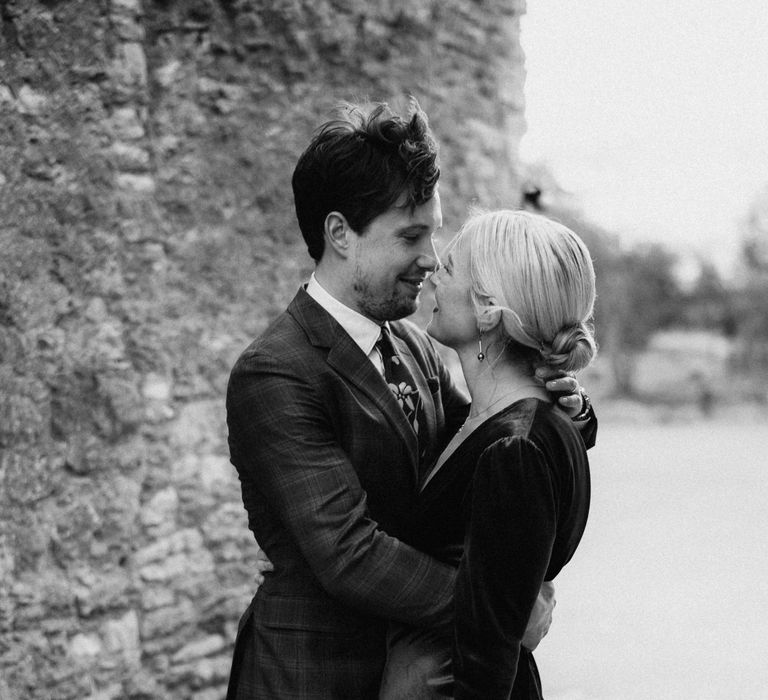 Bride wears her blonde hair pulled into low bun with pearl hair accessories whilst her groom wearing tartan suit wraps his arm around her