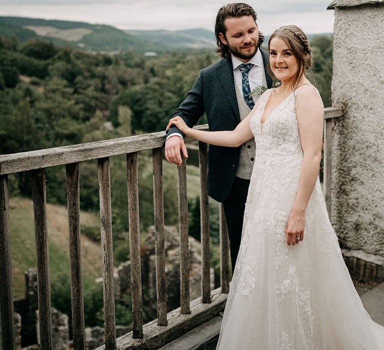 Bride & groom stand on balcony of castle outdoors in Scotland as bride wears lace wedding dress with floor-length veil in her brown hair