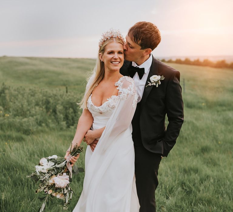 Groom in black tie kisses the bride during sunset with the bride in an embellished flower wedding dress and crown