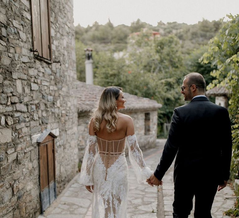 Bride wears lace fitted wedding dress with bell sleeves and low back whilst walking with her groom for beach bar wedding