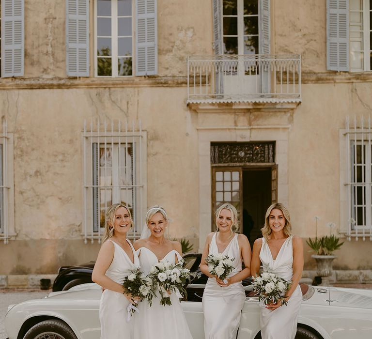 Bride stands in front of vintage car with her bridesmaids in white silk bridesmaid dresses with white rose bouquets