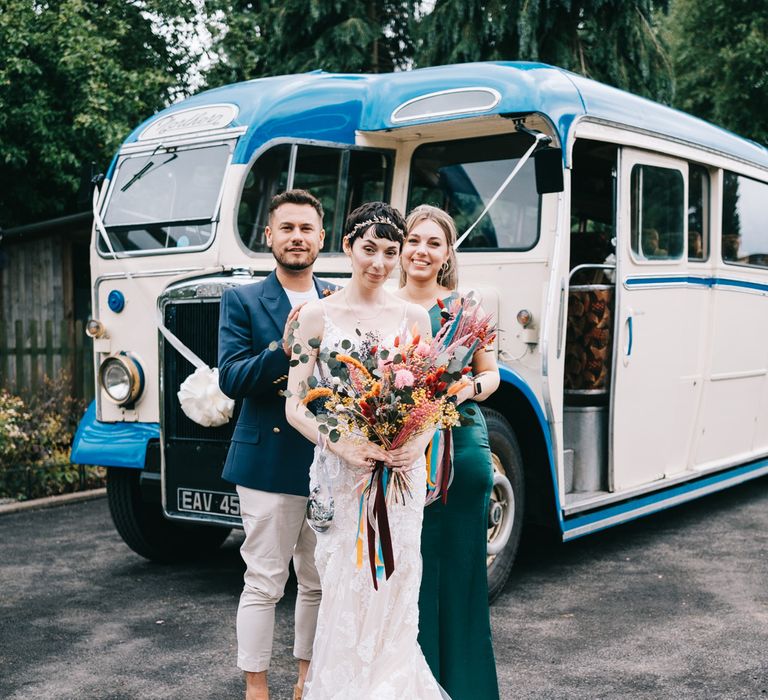 Bride holding bright dried floral bouquet stands with her wedding party in front of vintage bus