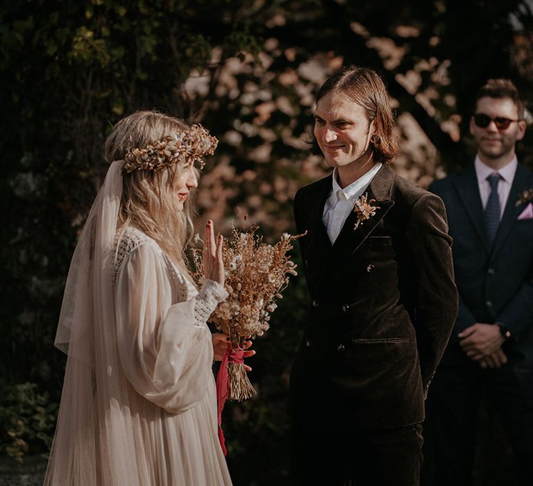 Bride wearing dried floral crown and bouquet stands in front of her groom in green suit during Pagan wedding 