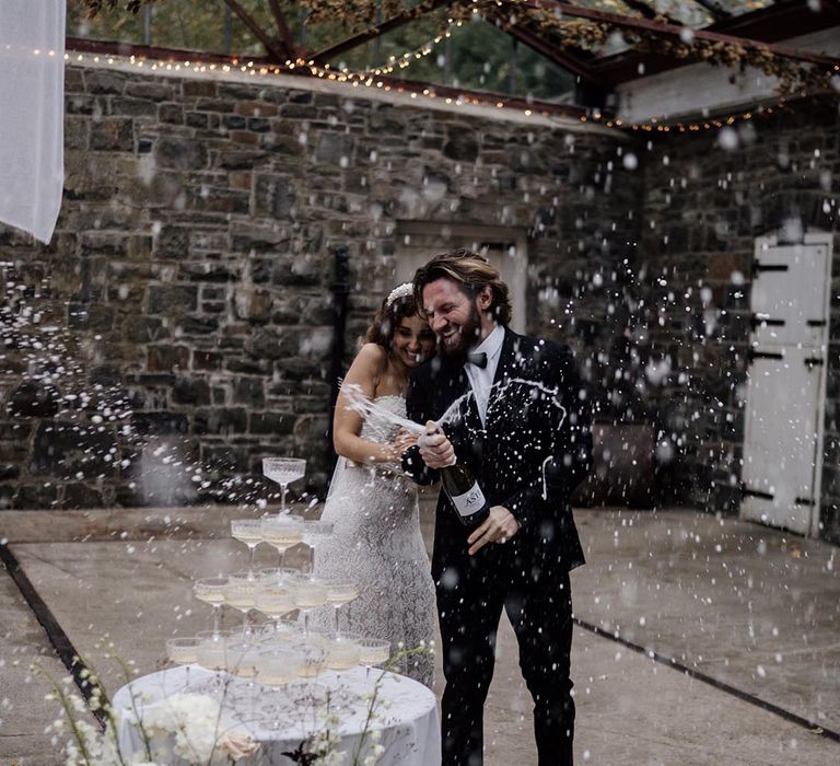 Bride in sleeveless lace detail wedding dress and groom in black tux popping Champagne 