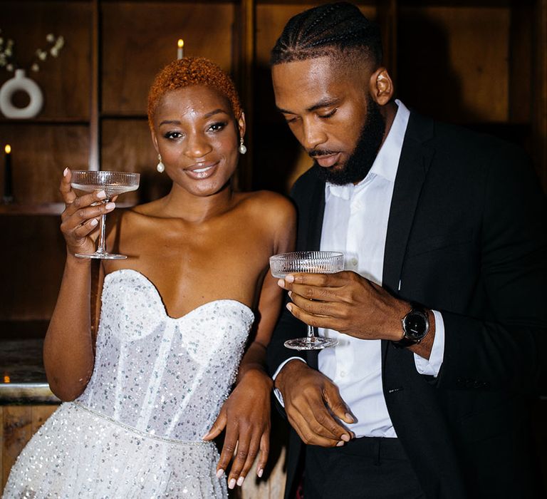 Bride in sparkly sweetheart strapless dress and leg slit holding a glass of champagne, standing with groom in skinny black suit