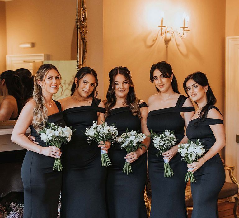 Bridesmaids in asymmetric one shoulder black bridesmaid dresses for the black tie wedding holding white flower bouquets 