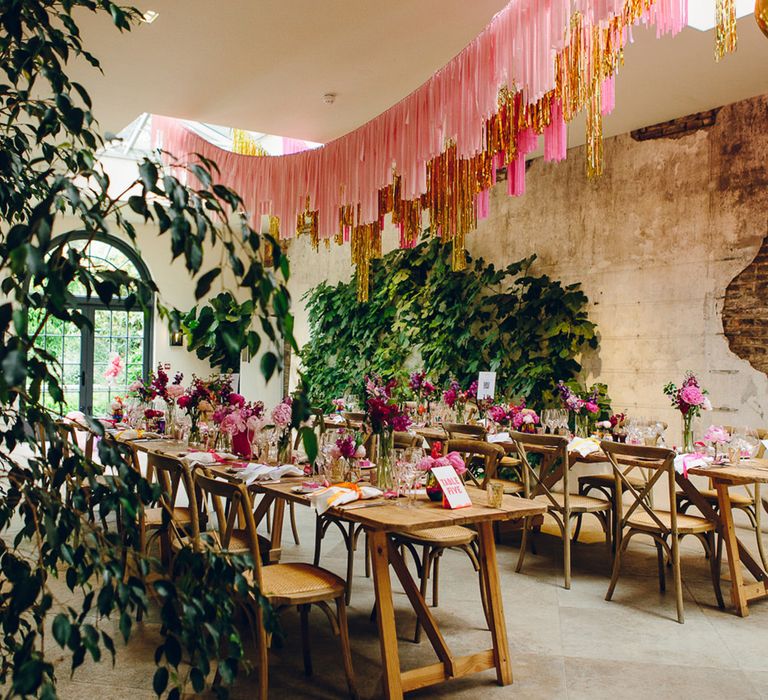 Neutral wedding tablescape with wooden tables, wooden chairs, large foliage decor, pops of pink, and pink and gold wedding streamers hanging above 