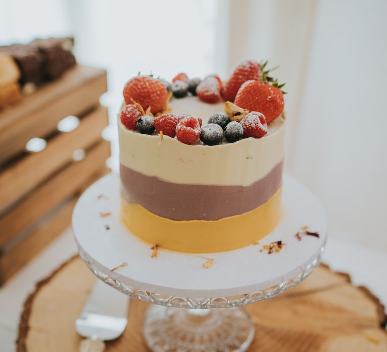 Single tier wedding cheesecake on wooden slate with cream, chocolate and orange icing and strawberries, blueberries and raspberries on the top 