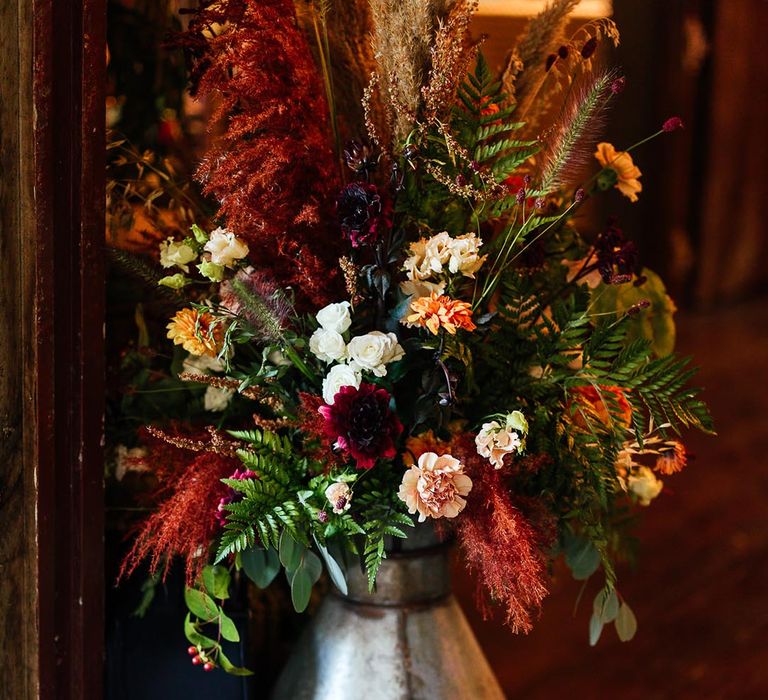 Boho autumnal wedding with orange and red flowers with fern leaves in a milk churn 