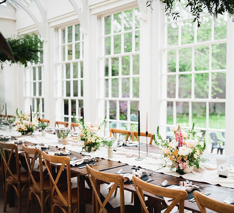 Reception room of The Orangery wedding venue with neutral off-white table runners, rose and foliage floral arrangements, pink and black tapered candles, black candlestick holders and suspended foliage decorations