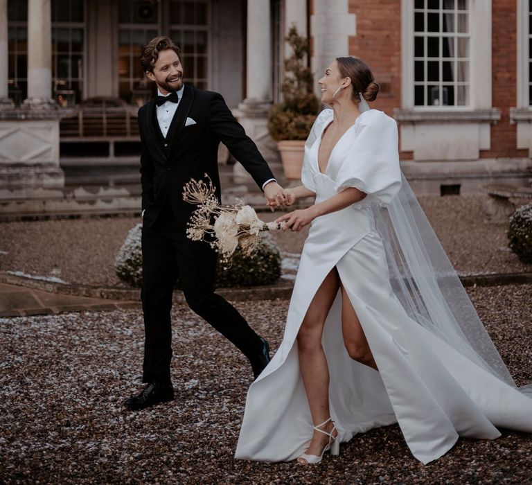 Bride in v neck puff sleeve wedding dress with front slit and church length veil and diamond drop earrings holding bouquet with large white roses and dried flowers and groom in classic black blazer and suit trousers with white shirt and white pocket square and black bowtie 