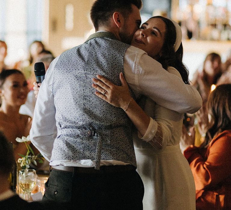 Bride holds a microphone as she delivers a bridal speech and hugs the groom in his waistcoat and shirt 