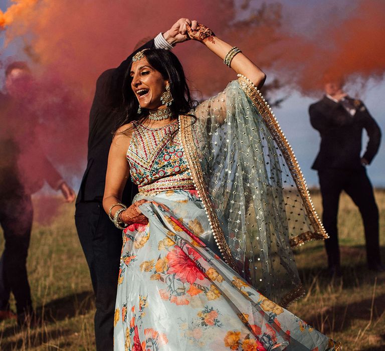 Bride in colourful floral lehenga dances outdoors during smoke-bomb portraits 