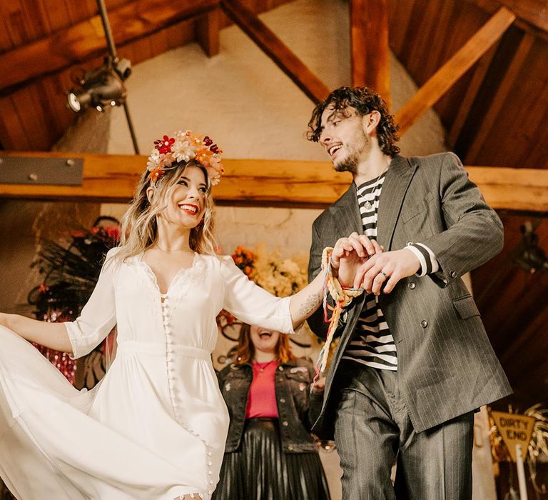 Groom in dark grey pinstripe suit and dr Martens boots and bride in three quarter length sheer sleeve wedding dress with button and cinched waist details, pink bridal flower crown and pink fluffy open-toe strappy heels doing jumping the broom wedding tradition