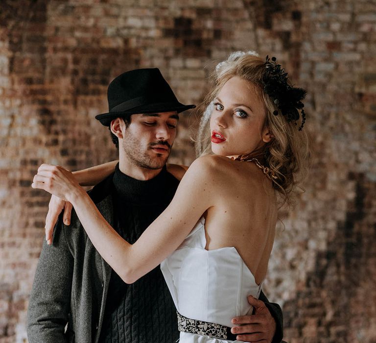 Bride in sculpted bandeau top wedding dress with full ballgown skirt and patterned black belt embracing groom in dark grey jacket, black ribbed turtle neck, black trousers and a black fedora hat 