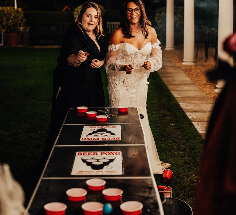 The bride in a beaded floral wedding dress plays beer pong on black shiny table 