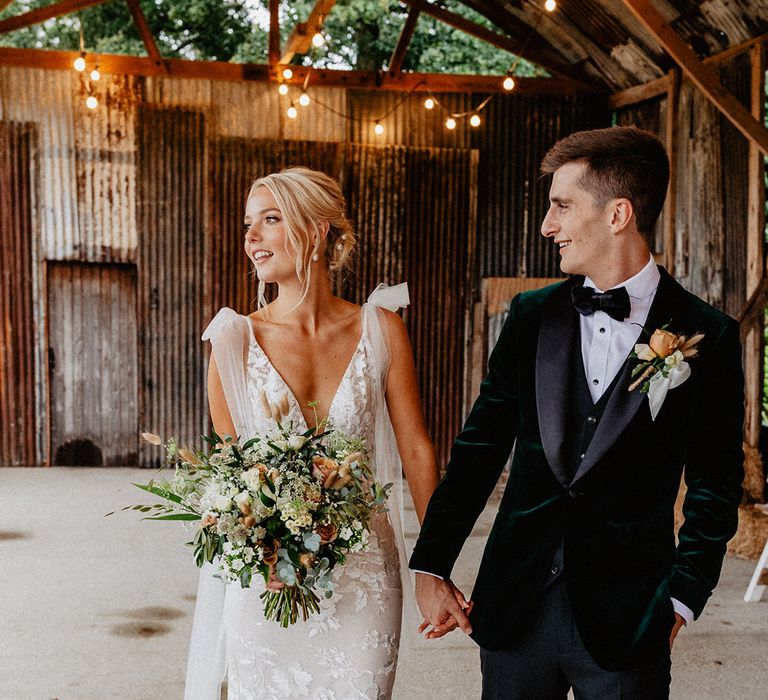 The groom in velvet suit jacket with rose buttonhole with ribbon walks along with the bride in her Made With Love wedding dress 