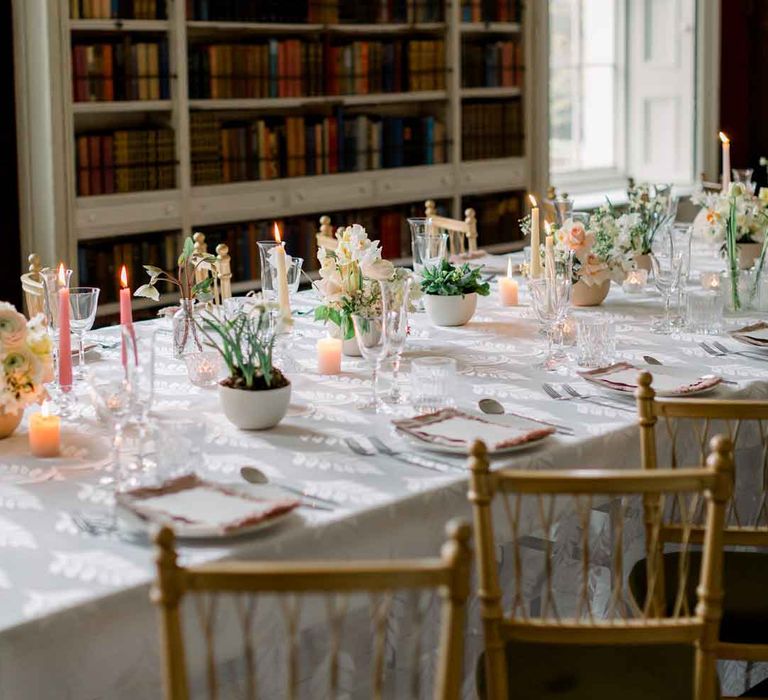 CBride wearing layered, sculpted off the shoulder wedding dress and groom in navy blue suit with yellow tie sat at classic wedding tablescape with white tablecloth, pink tapered candles and spring florals at St Giles House