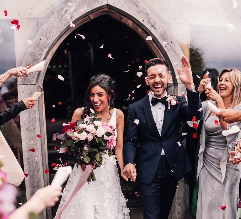 Bride in floral appliqué Wedding dress walking with the groom in black tie out from their ceremony with petal confetti moment 