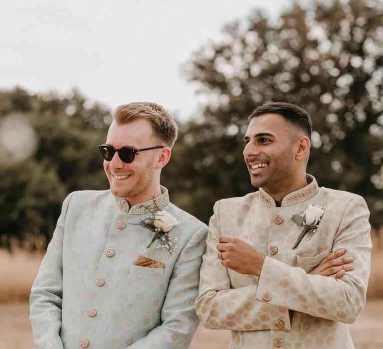 Groom in mint green Indian Sherwani with white garden rose and foliage boutonniere and grooms sunglasses standing with groom in cream and gold Indian sherwani and white garden rose boutonniere at Gujarati wedding ceremony at High Billinghurst Farm