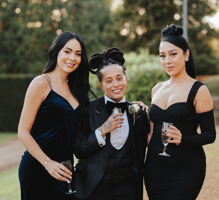 Bride in black snakeskin print tuxedo with black bowtie and white rose boutonniere standing with bridesmaids in mismatched black bridesmaid dresses 