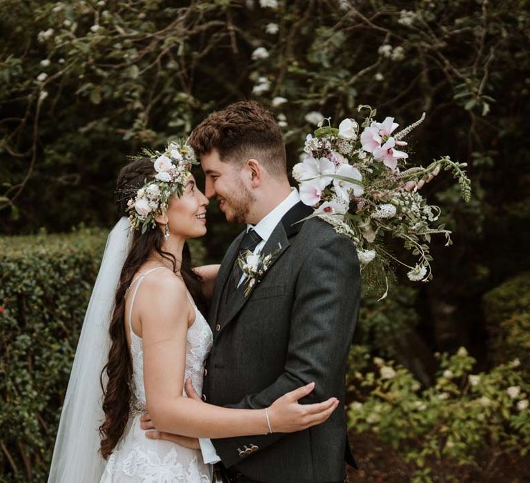 Bride in 3d applique sheer overlay wedding dress with white garden rose flower crown and cathedral-length veil embracing groom in dark grey blazer with tartan tie and white garden rose boutonniere 