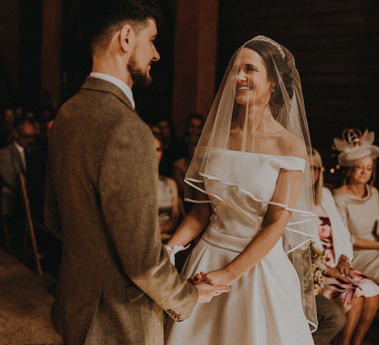 The bride in an off the shoulder wedding dress and veil smiles facing the groom for their wedding ceremony 