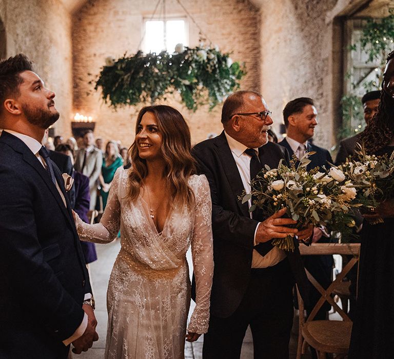 Bride in sparkly winter wedding dress meeting the groom at the end of the aisle in a navy suit for their barn wedding 