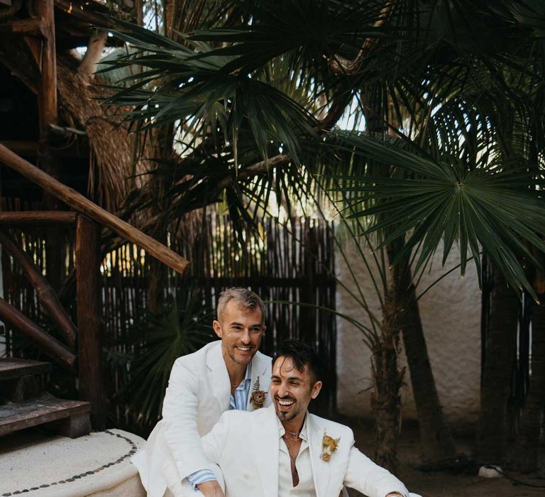 Groom sitting on stairs of hotel wedding venue wearing white blazer with dusky orange garden rose and dried flower boutonniere, white shirt, white suit trousers and silver jewellery with groom in white mens wedding suit,  light blue and white striped shirt, dusky orange garden rose and dried flower boutonniere, white suit trousers and gold jewellery 