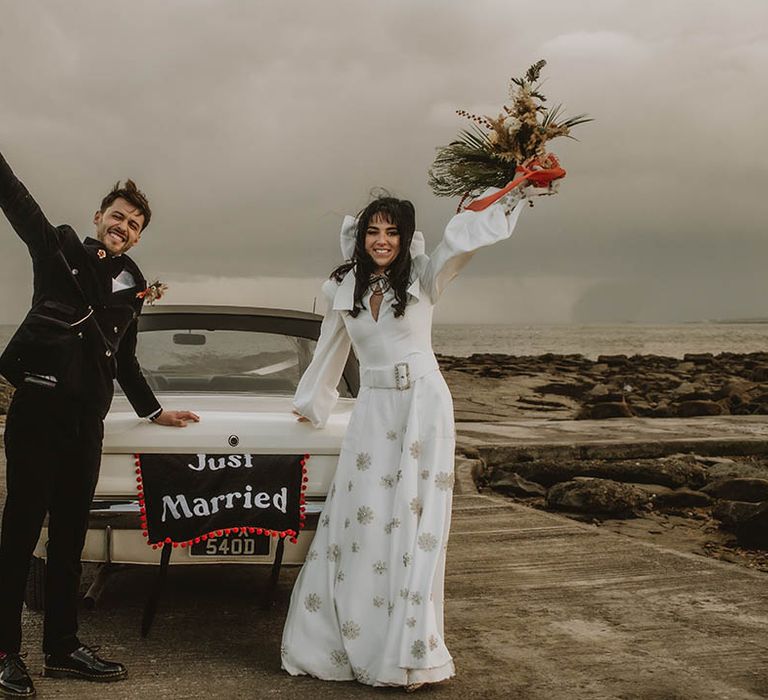 Bride in custom sparkly Bowen Dryden bridal jumpsuit with the groom in front of their wedding car decorated with wedding banner 