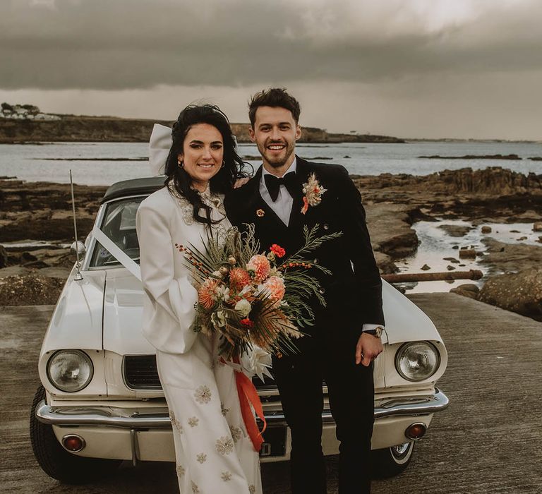 Bride in sparkly Bowen Dryden jumpsuit and large white hair bow accessory with groom in black velvet suit 