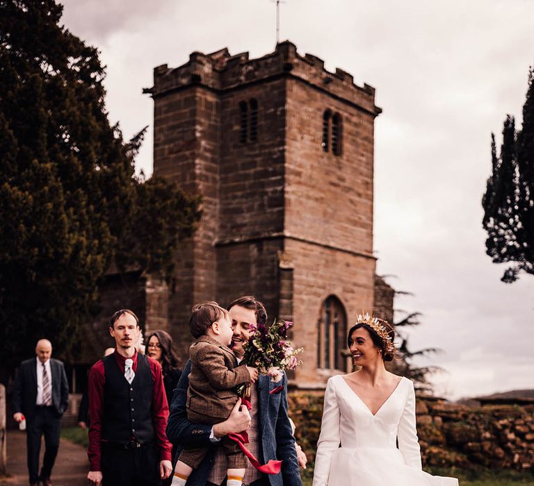 Groom in navy three piece suit with red tie carrying their toddler in a brown suit walking with the bride in a traditional princess style wedding dress