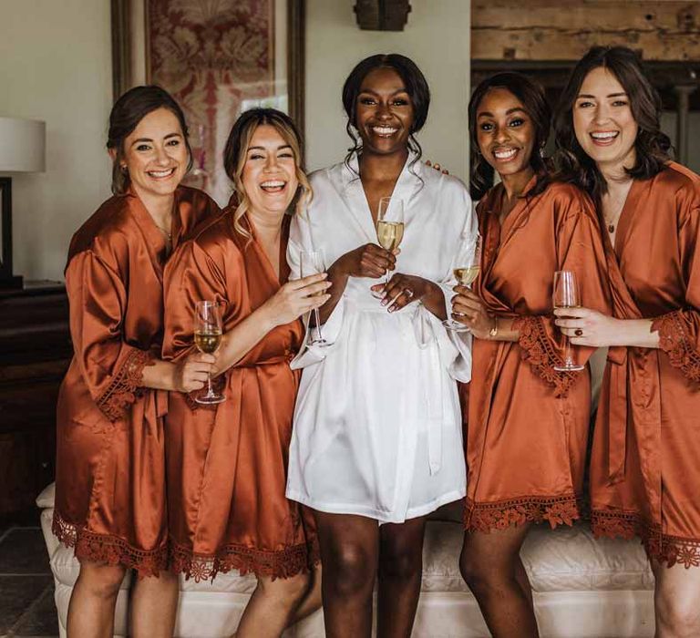 Bride in white satin getting ready robe with bridal party wearing burnt orange getting ready robes at Lains Barn wedding venue 