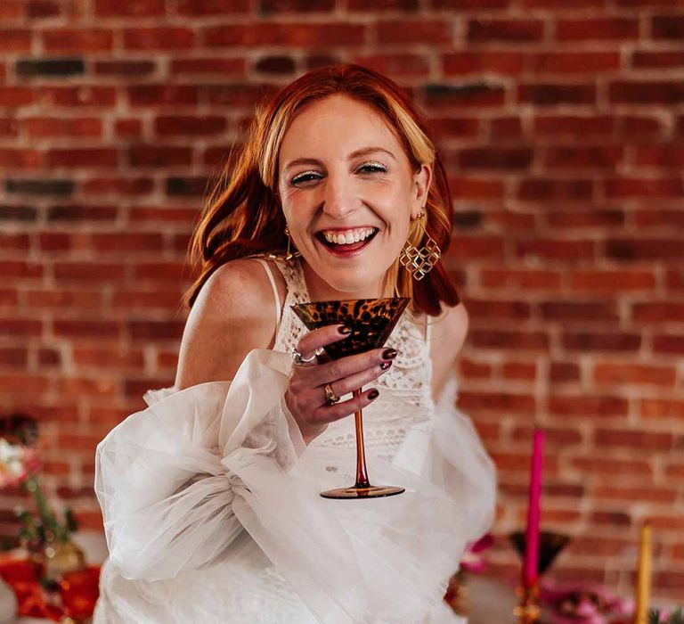 Bride in 70s-inspired boatneck short lace wedding dress with detachable tulle puff sleeves and platform gold wedding heels sipping from leopard print cocktail glass holding colourful mixed flower bridal bouquet 