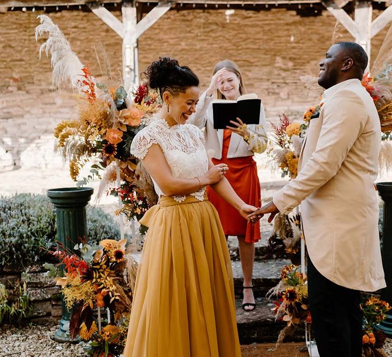 Groom in cream morning suit with forest green grooms waistcoat and trousers, forest green pocket square and orange boutonniere standing and holding hands with bride in white mesh lace bridal crop top and yellow wedding skirt