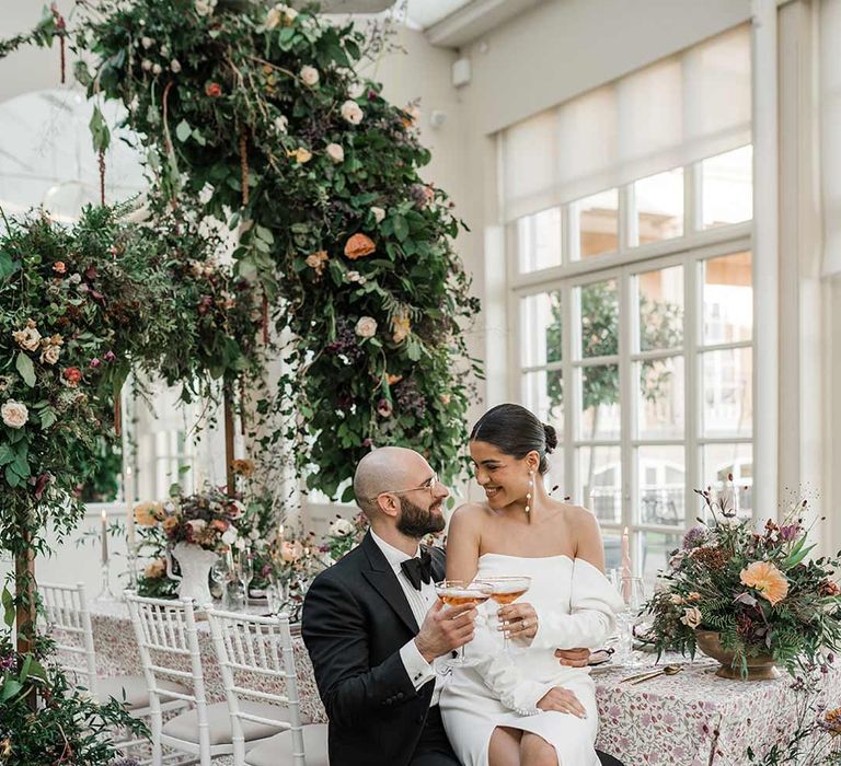 Groom in classic black tuxedo with bowtie and wildflower boutonniere with bride on his lap wearing long sleeve off the shoulder wedding dress with pearl button detailing and a side slit holding cocktail sitting by botanical wedding tablescape with patterned floral tablecloth, twisted tapered candles, art deco vases with dried wildflower wedding centrepieces surrounded by suspended wildflower and foliage arrangements in large gold vases at Buxted Park